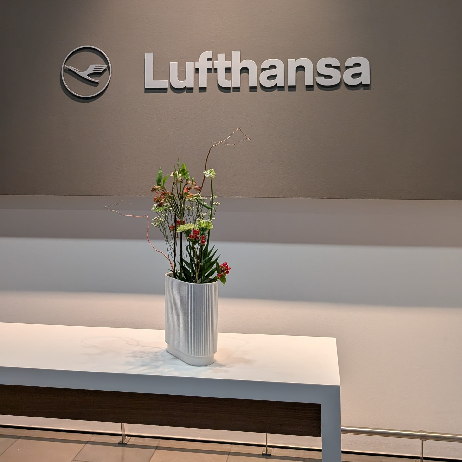Nahaufnahme eines weißen Empfangstresens in dem Eingangsbereich des Lufthansa First Class Terminals in Frankfurt. Auf dem Tresen steht eine elegante, weiße Vase mit einem filigranen Blumenarrangement aus grünen Zweigen und kleinen roten Akzenten. Im Hintergrund prangt an einer grauen Wand das silberne Lufthansa-Logo sowie der Lufthansa-Schriftzug unter einer sanften Spot-Beleuchtung.