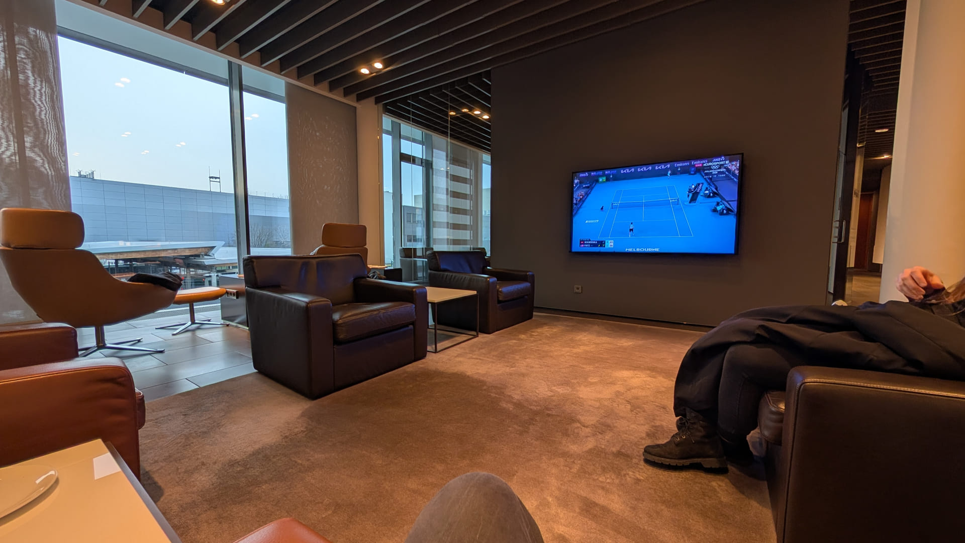 Ein luxuriöser Loungebereich im Lufthansa First Class Terminal. Der Raum ist mit schweren, dunklen Ledersesseln auf einem hellbraunen Teppich ausgestattet. An einer dunkelgrauen Wand hängt ein großer Flachbildfernseher, auf dem ein Tennisspiel übertragen wird. Große Fensterfronten im Hintergrund lassen Tageslicht herein und bieten einen Blick auf das Flughafengelände. Die Atmosphäre ist ruhig und exklusiv.